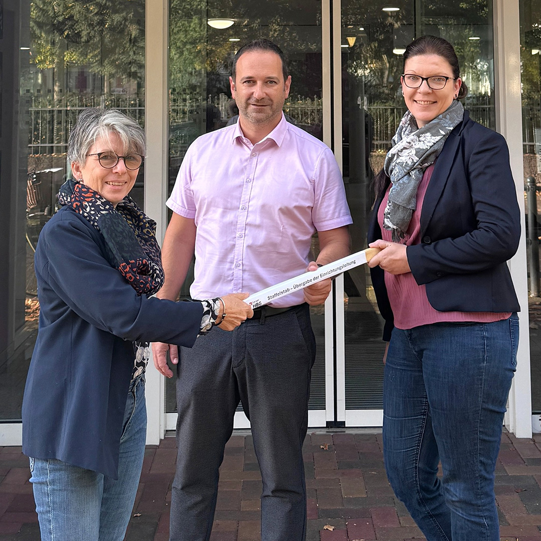 Drei Personen stehen vor dem Eingang des Pflegezentrums am Hofwiesenpark in Gera und halten einen beschrifteten Staffelstab zur Übergabe der Einrichtungsleitung.