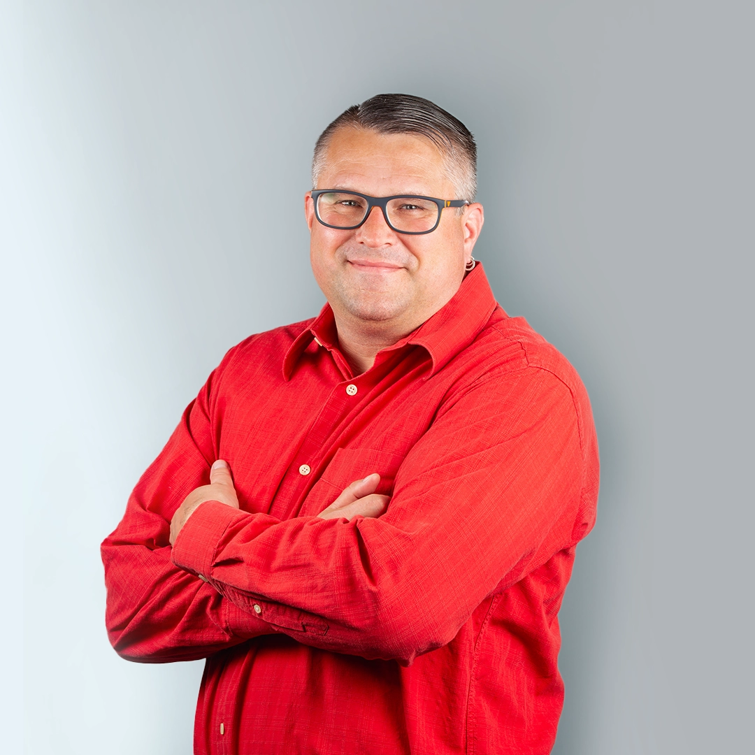Porträt eines Mannes mit Brille und rotem Hemd, verschränkten Armen und freundlichem Blick.