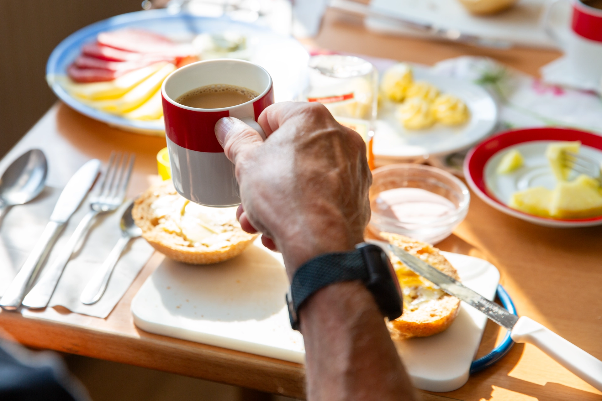 Hand hält eine Kaffeetasse über gedecktem Frühstückstisch mit Brötchen und Aufschnitt