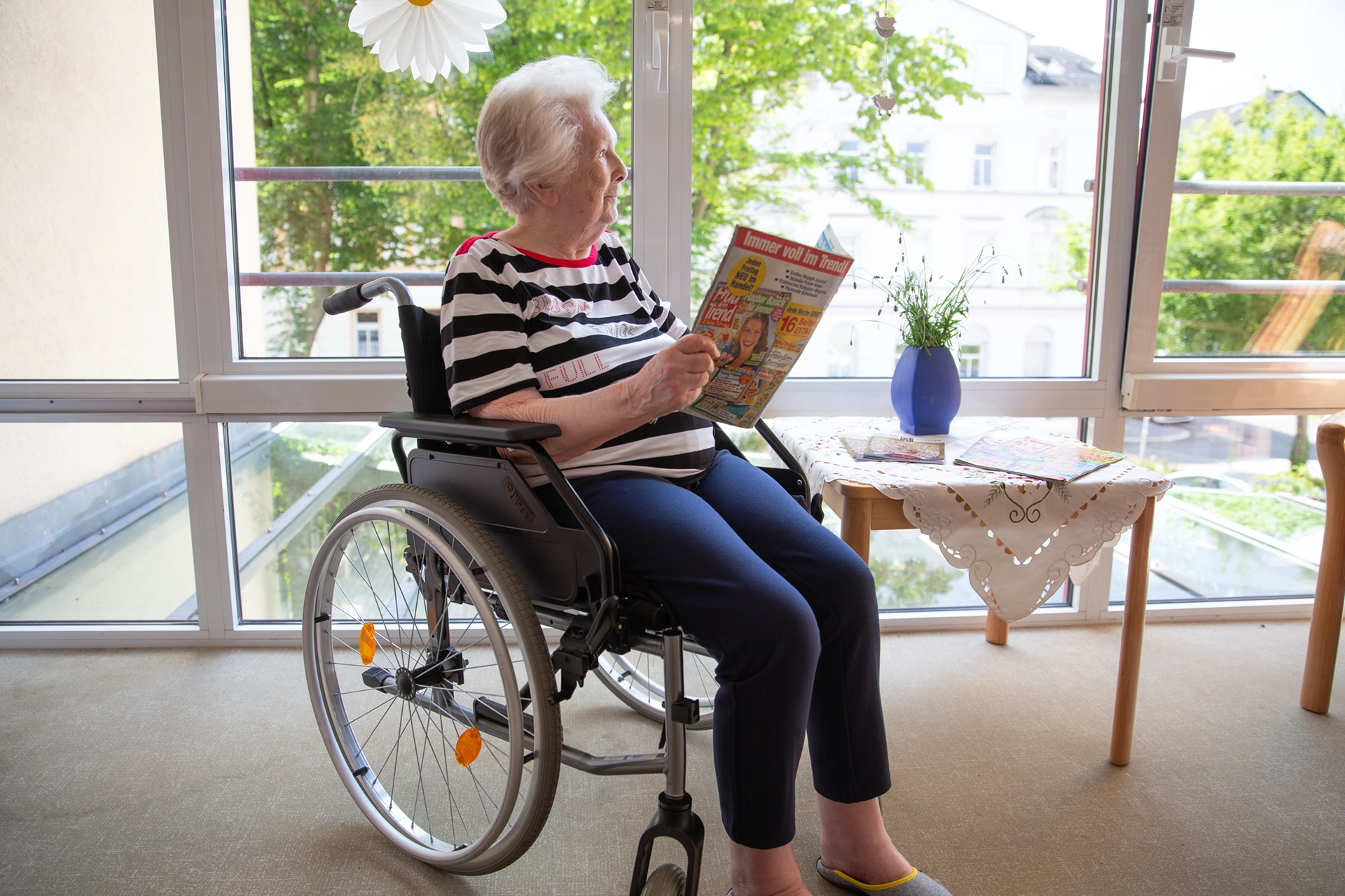Seniorin im Rollstuhl liest am Fenster eine Zeitschrift.
