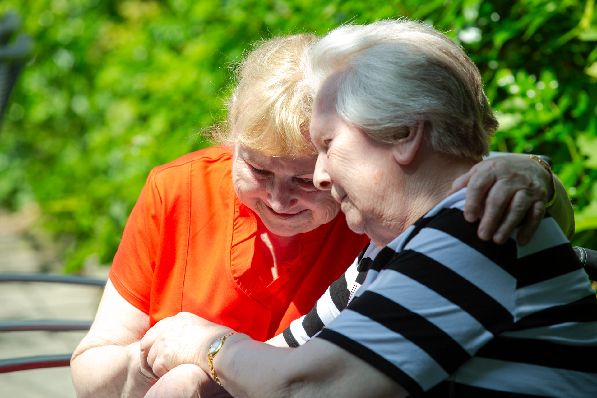 Zwei Seniorinnen umarmen sich herzlich im Garten.