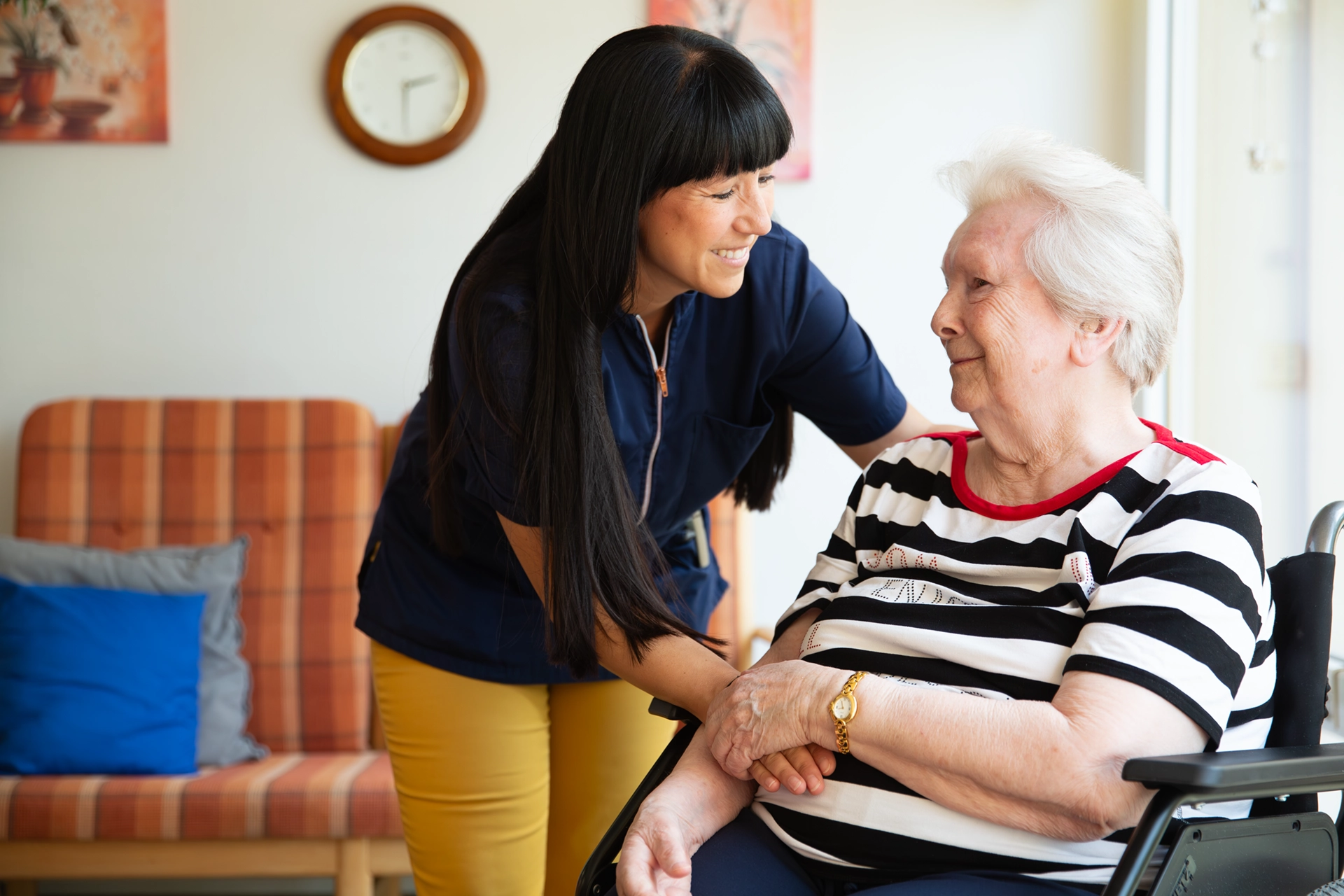 Pflegekraft beugt sich zu lächelnder Seniorin im Rollstuhl.
