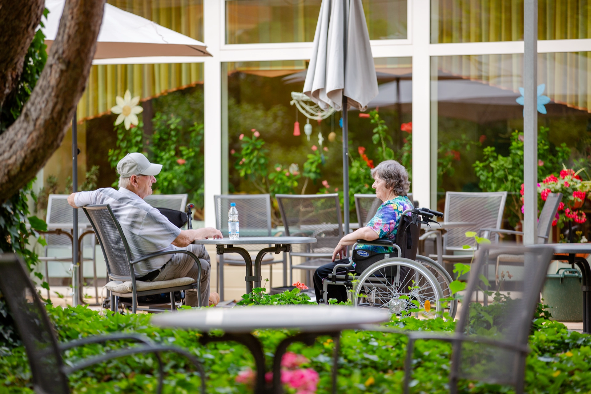 Zwei ältere Menschen sitzen sich auf der Terrasse gegenüber, einer im Rollstuhl.