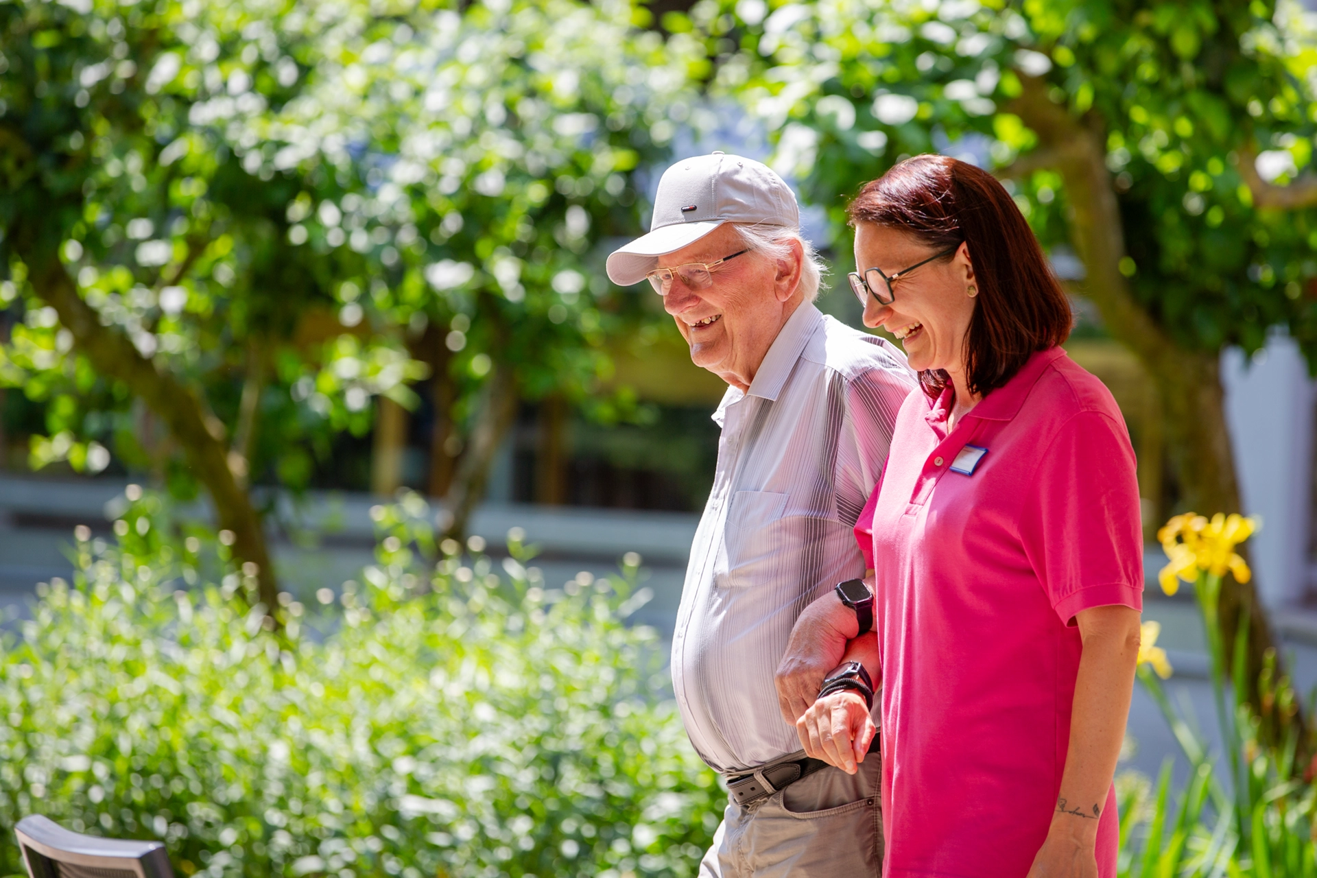 Eine Pflegekraft geht mit einem älteren Herrn lachend durch den Garten.