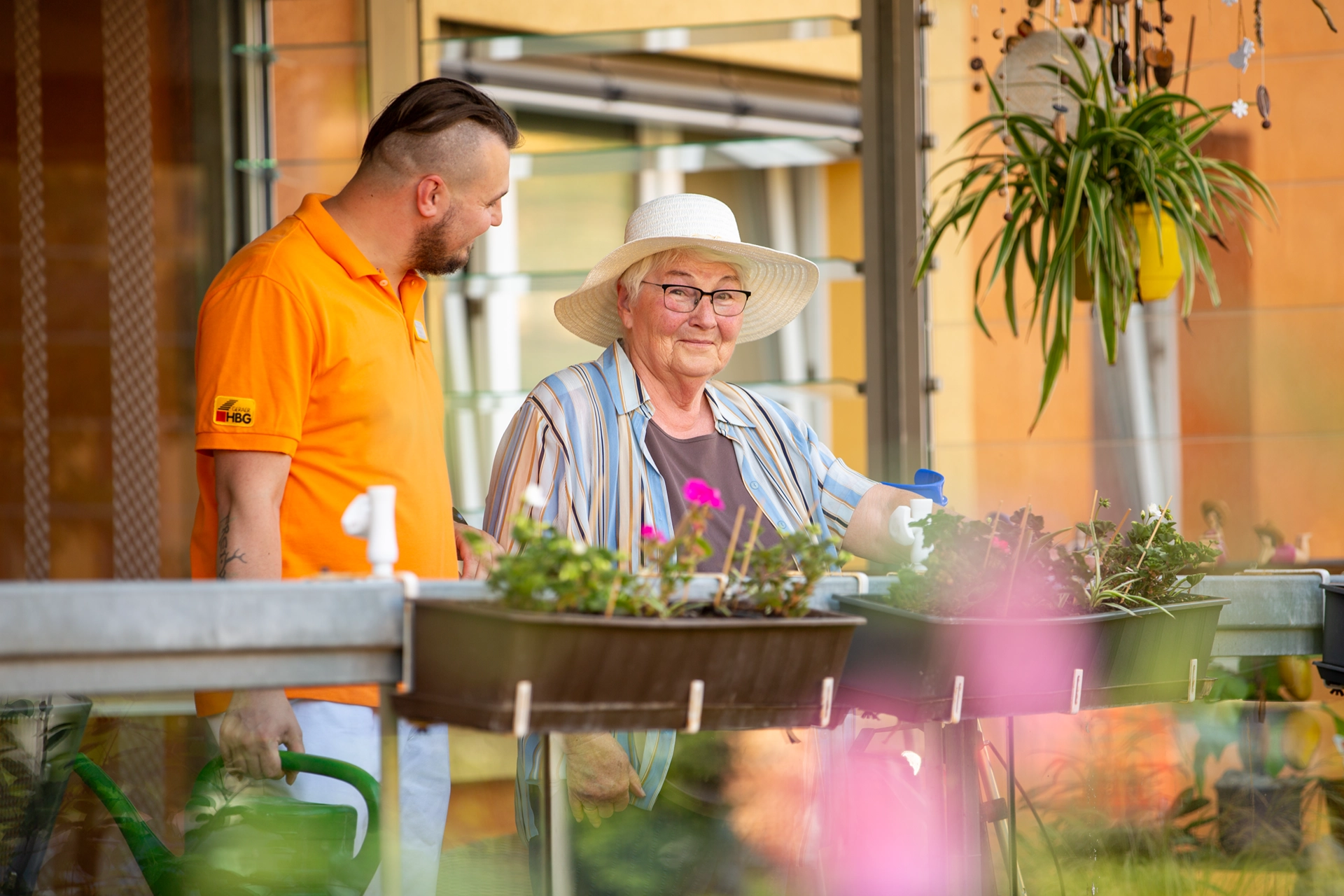 Bewohnerin mit Sonnenhut pflegt Blumen auf dem Balkon, begleitet von einem Pfleger.