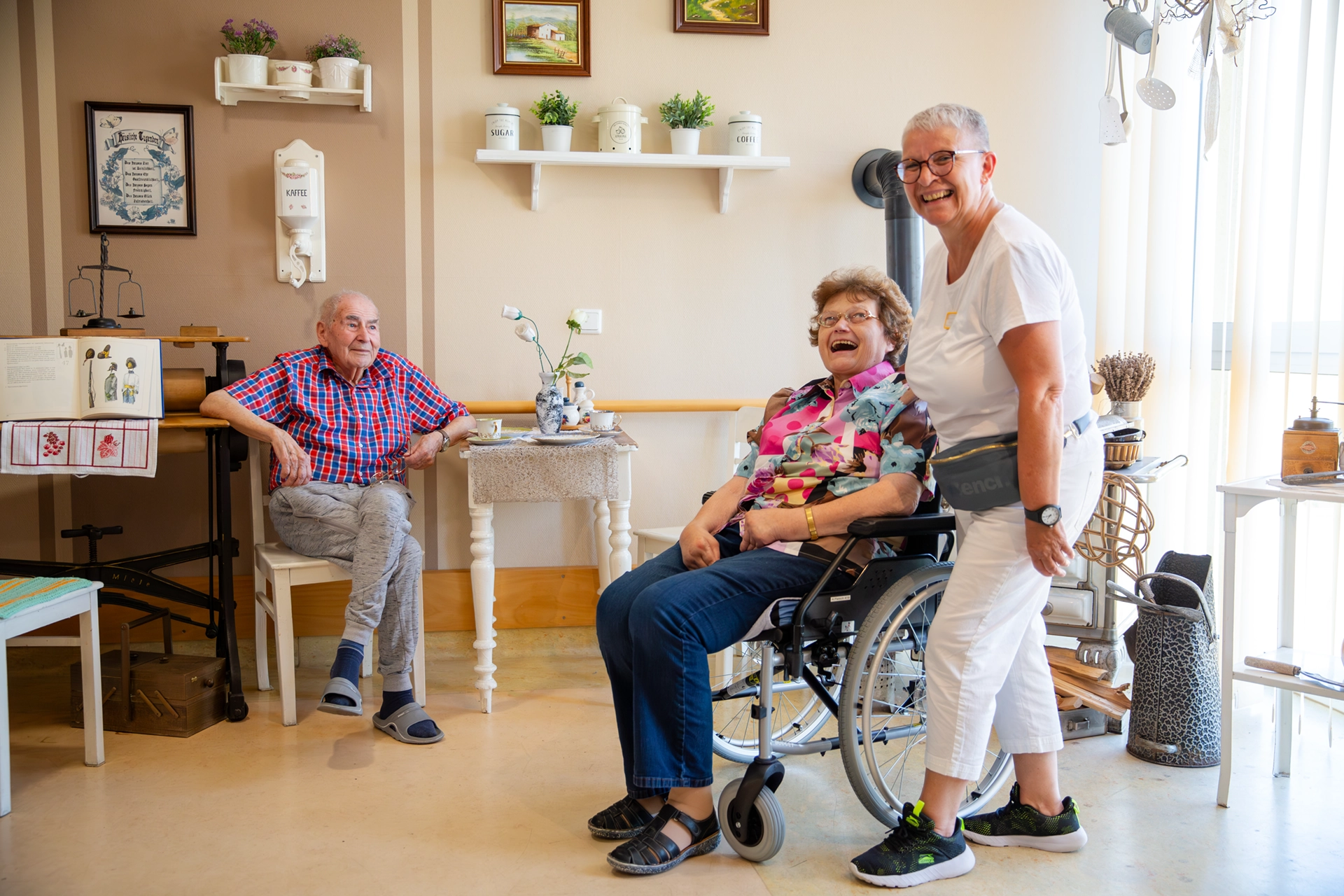 Drei Personen lachen gemeinsam in einem gemütlichen Raum mit Alltagsgegenständen.