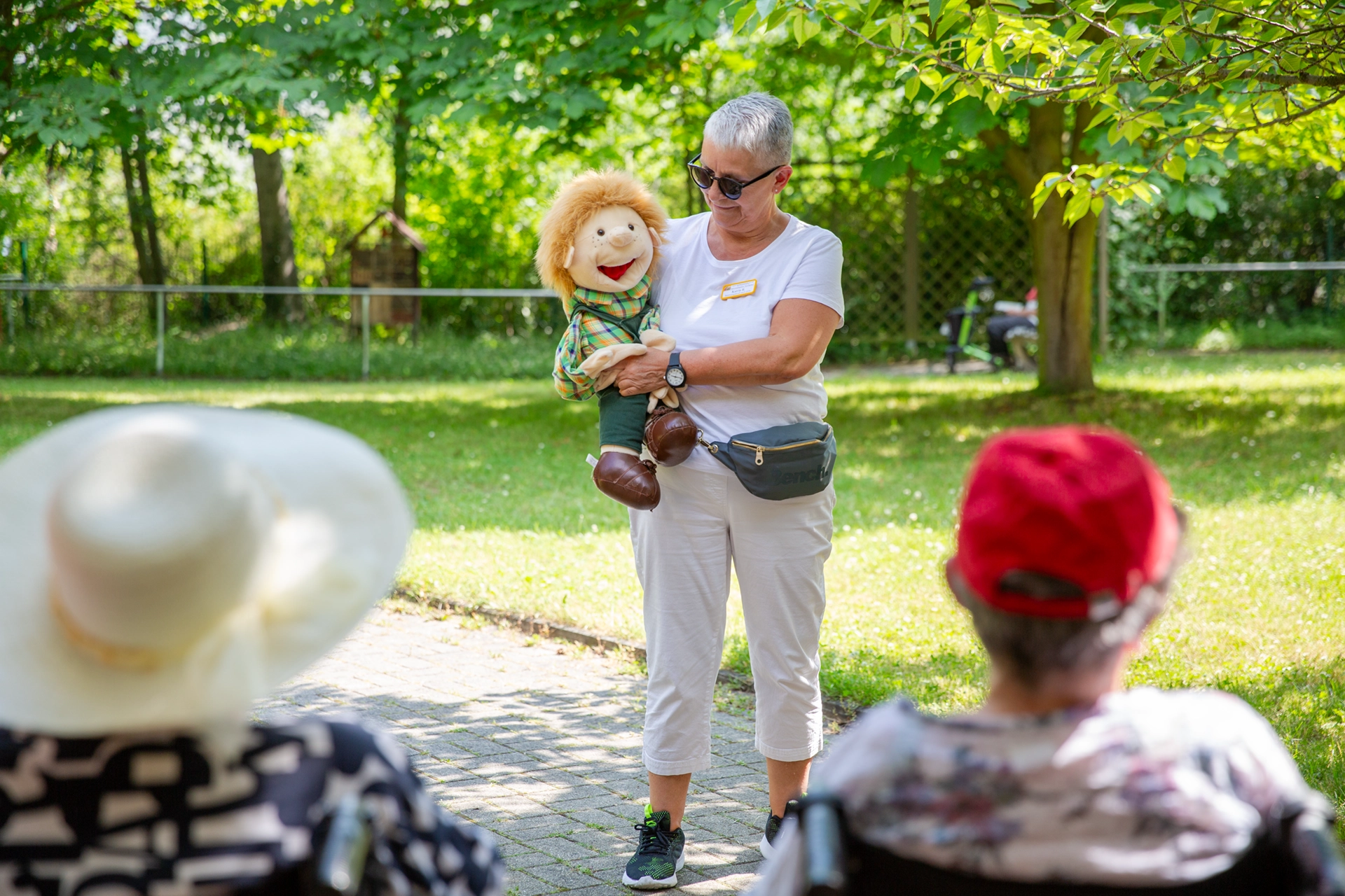Frau mit Handpuppe spricht mit älteren Menschen im Garten des Pflegeheims Kurt Neubert.