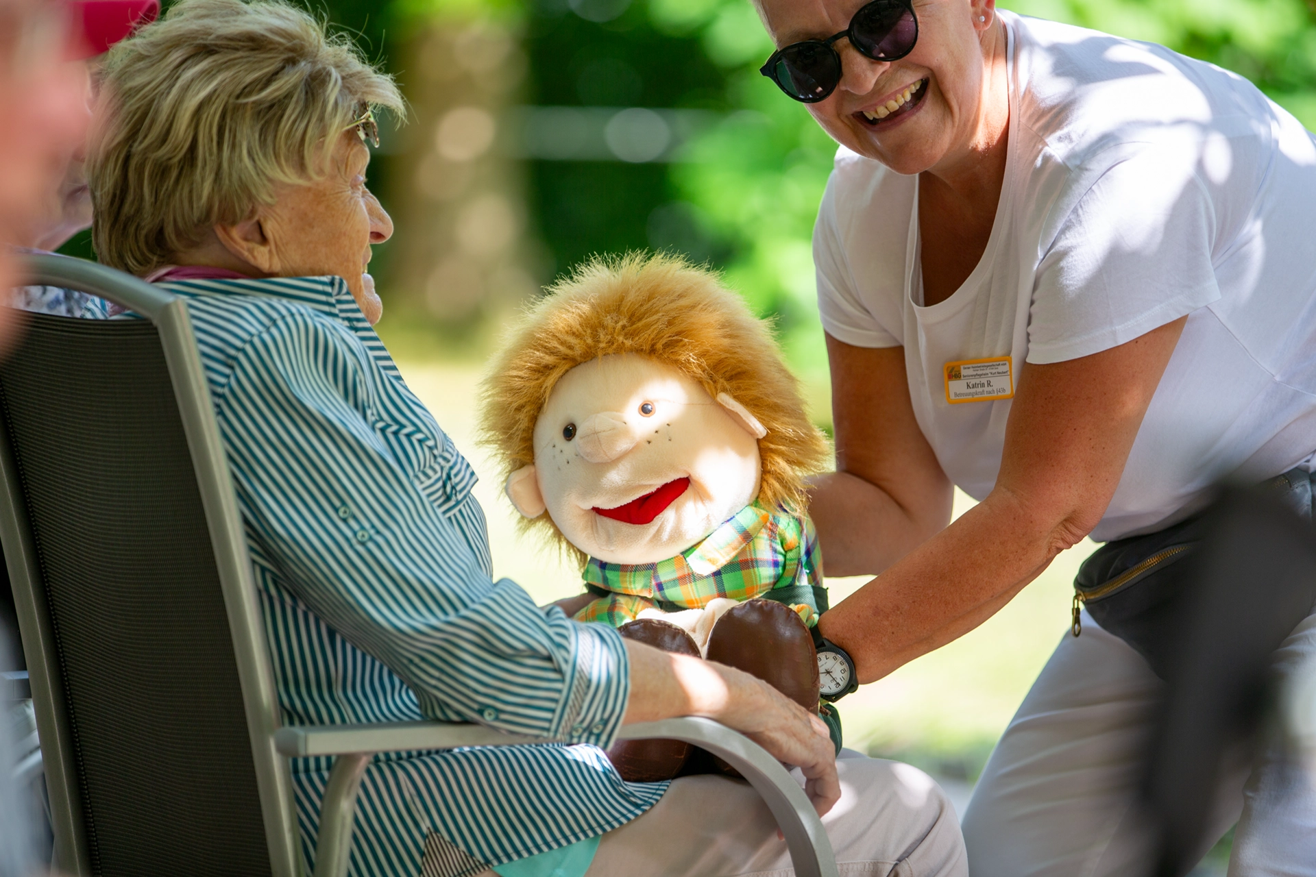 Ältere Frau interagiert lachend mit einer Handpuppe, gehalten von einer Mitarbeiterin.