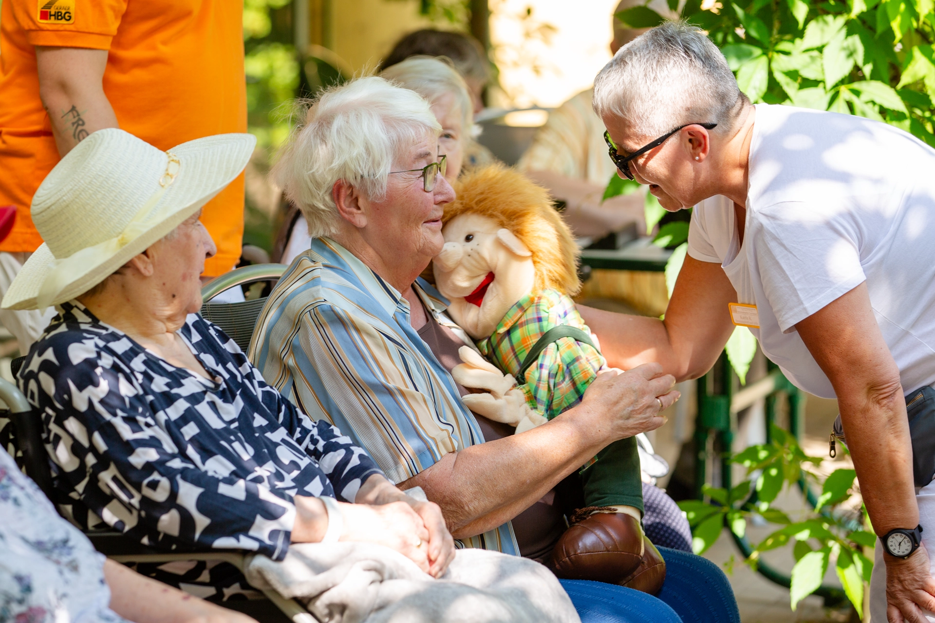 Seniorin hält eine Puppe auf dem Schoß, Pflegerin spricht sie liebevoll an