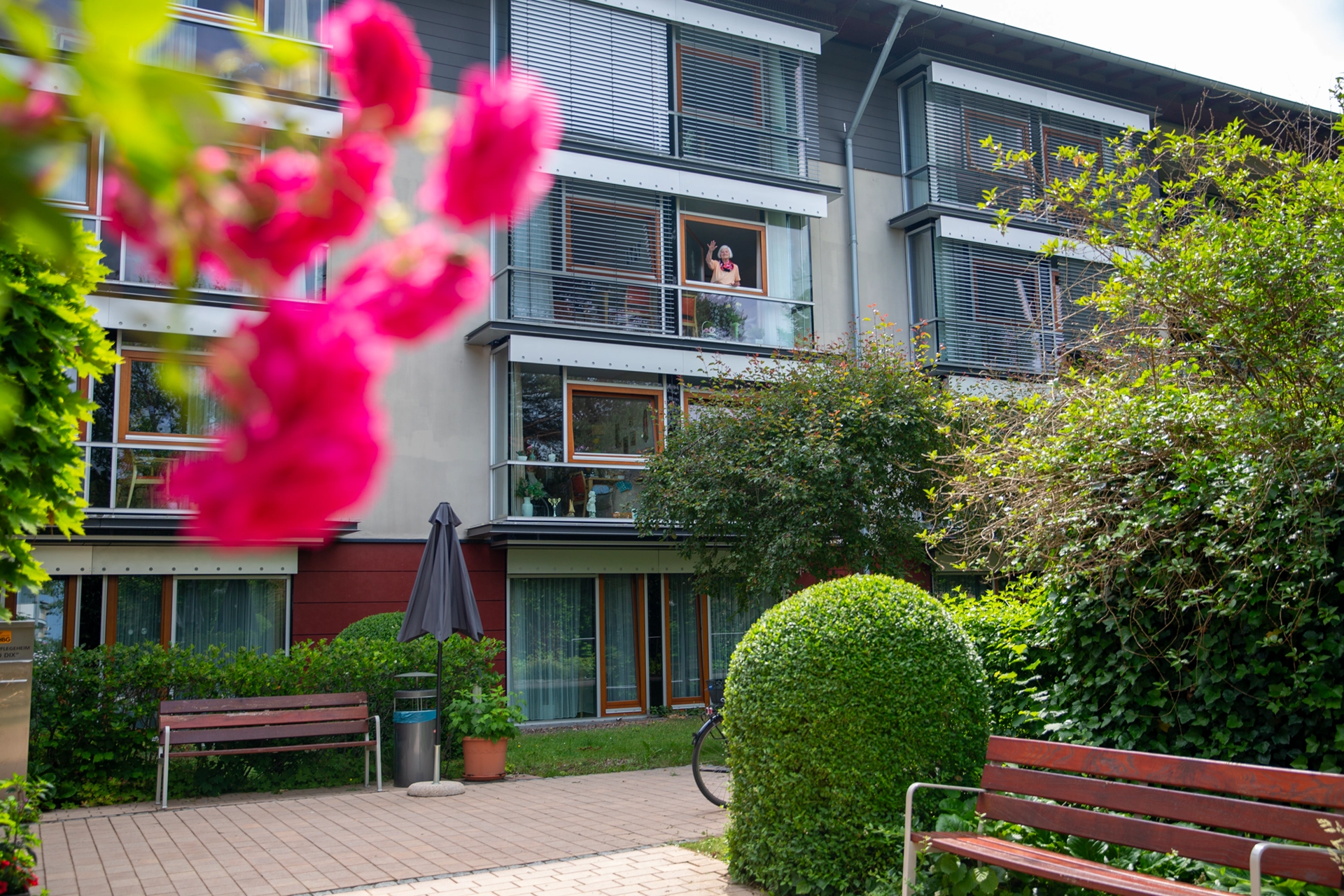 Außenansicht des Pflegeheims mit Garten, Büschen und einer winkenden Bewohnerin am Fenster