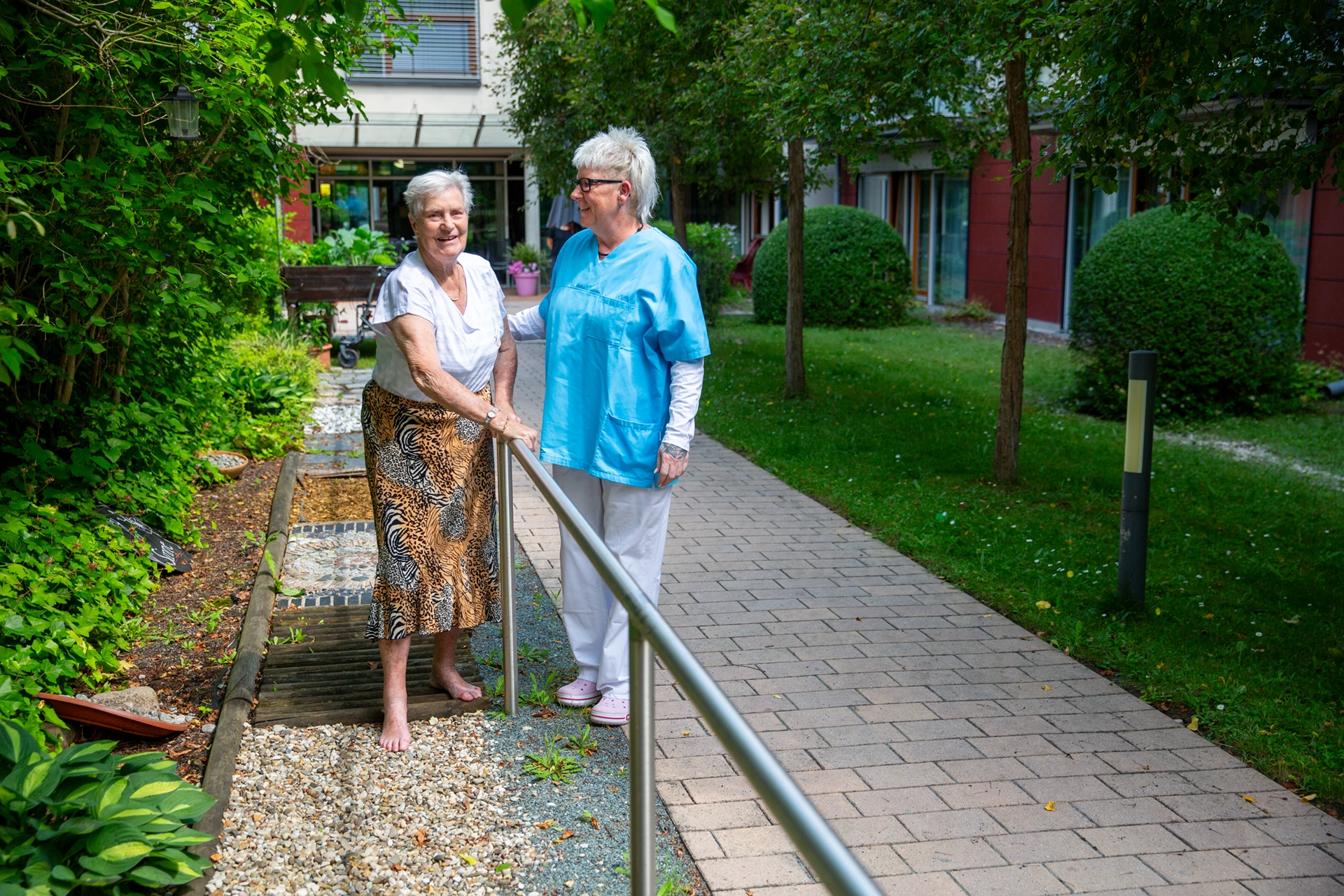 Zwei Frauen auf einem Barfußpfad im Garten, eine davon ist eine Pflegekraft