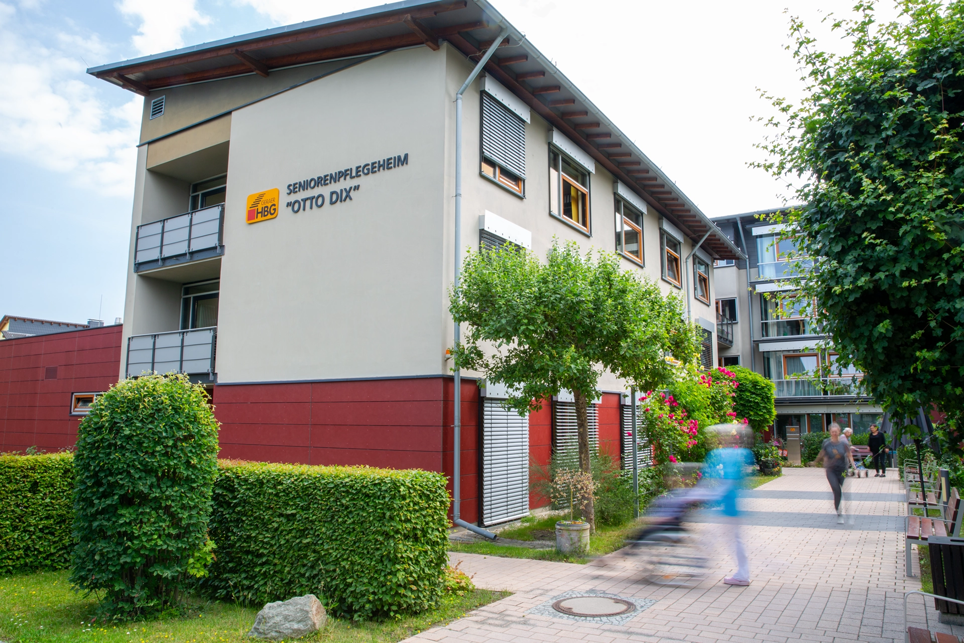 Haupteingang des Pflegeheims Otto Dix mit HBG-Logo und Menschen im Vordergrund