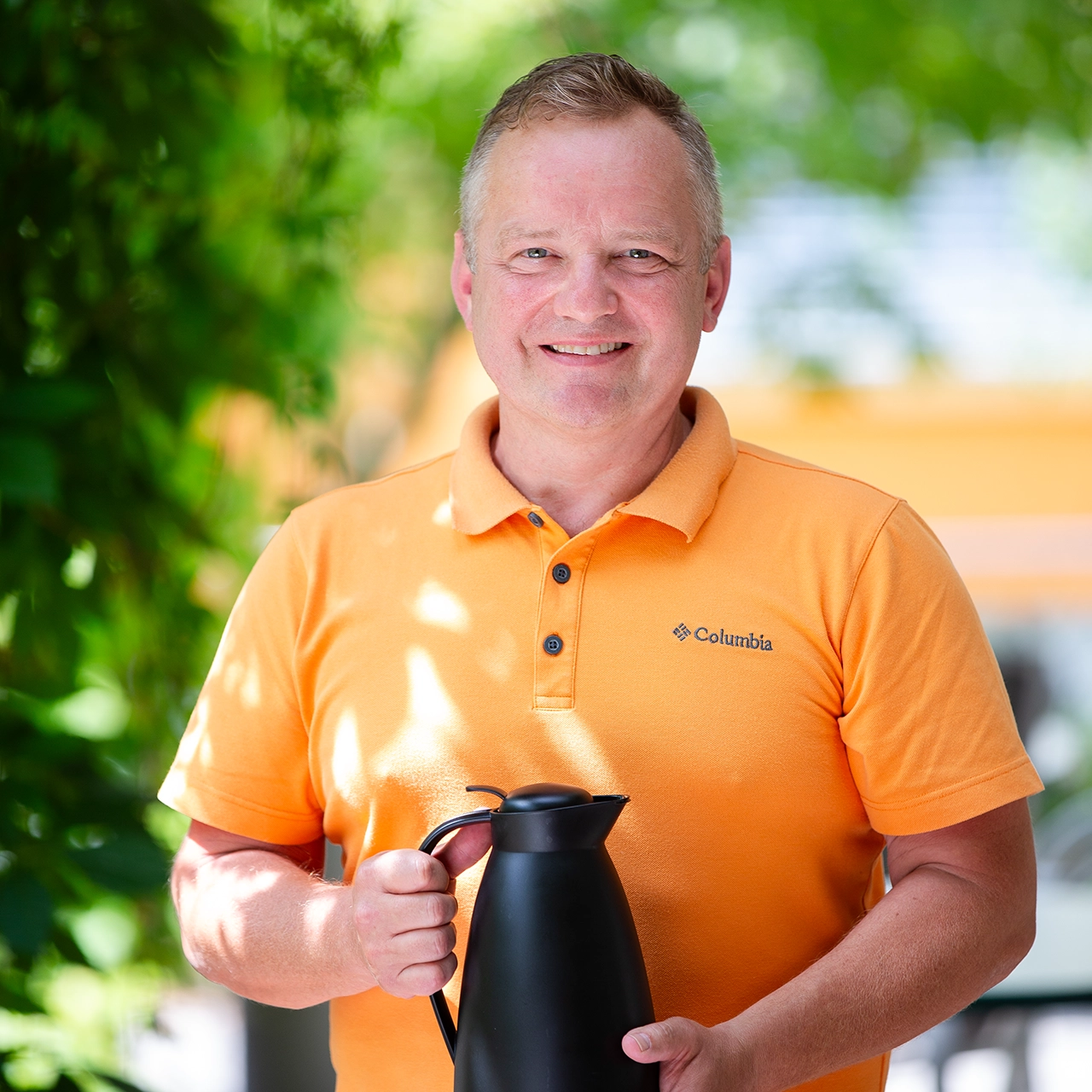 Mitarbeiter in orangefarbenem Poloshirt lächelt unter Bäumen.
