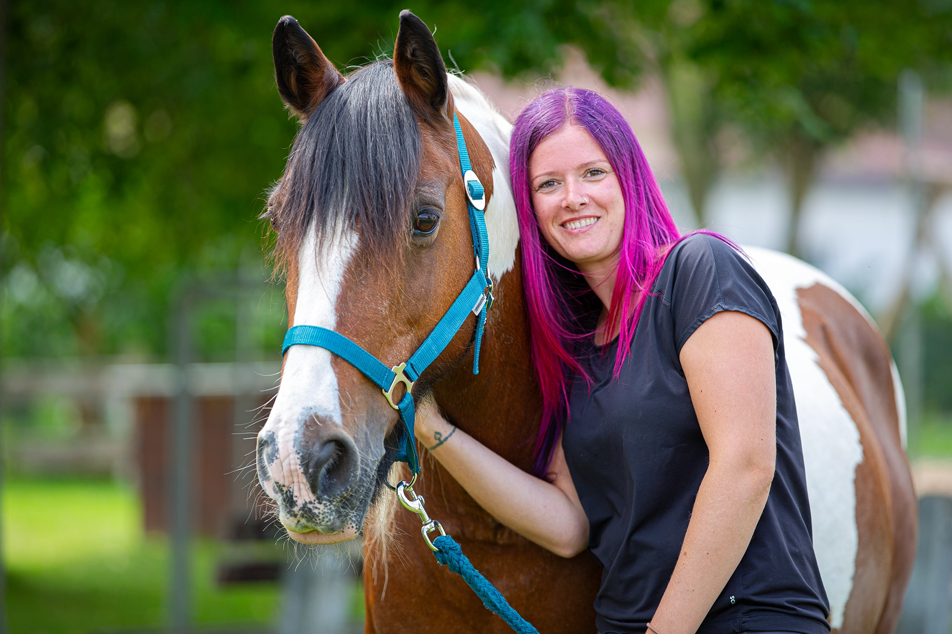 Frau mit pink-violetten Haaren steht lächelnd neben einem braun-weißen Pferd mit blauem Halfter.