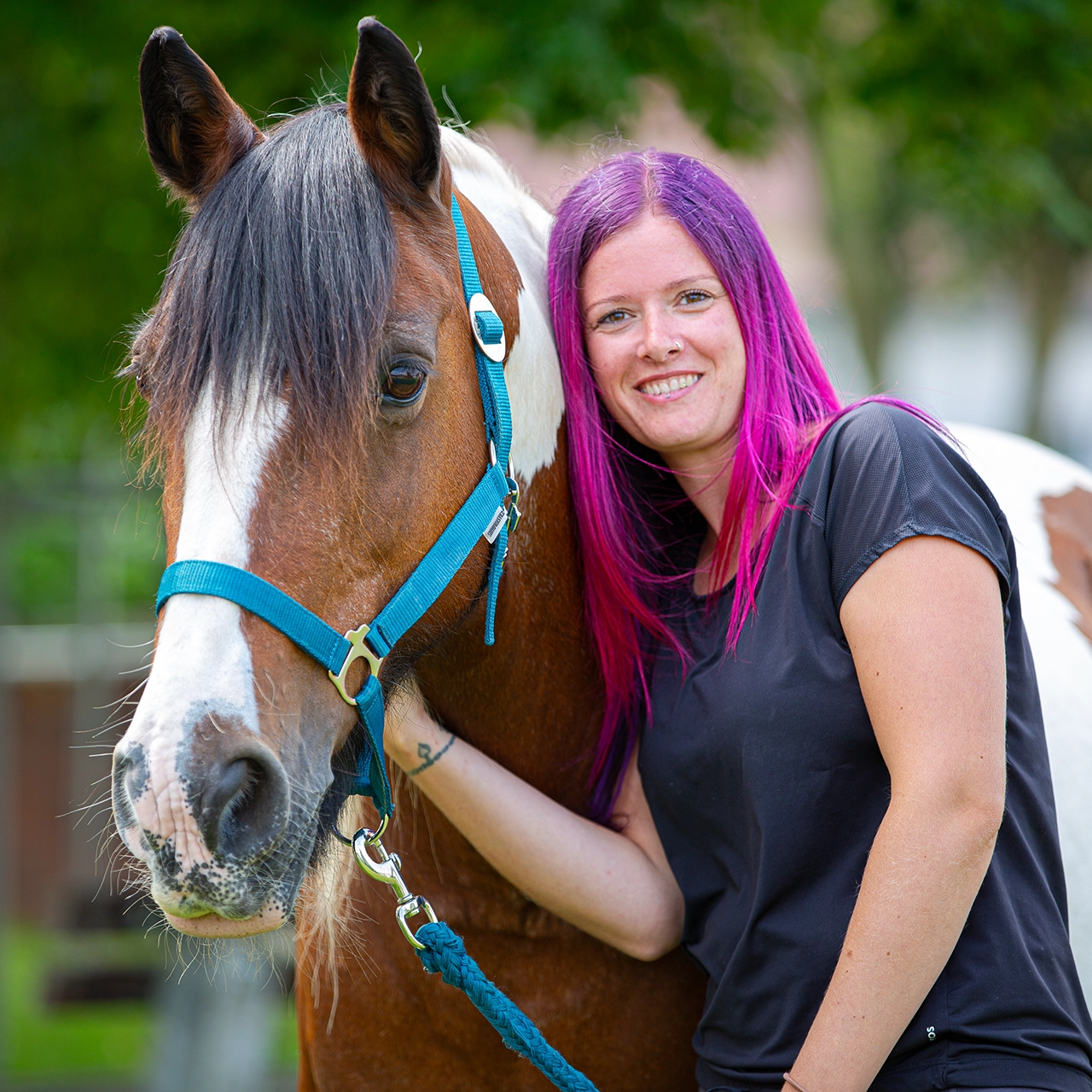 Frau mit pink-violetten Haaren steht lächelnd neben einem braun-weißen Pferd mit blauem Halfter.